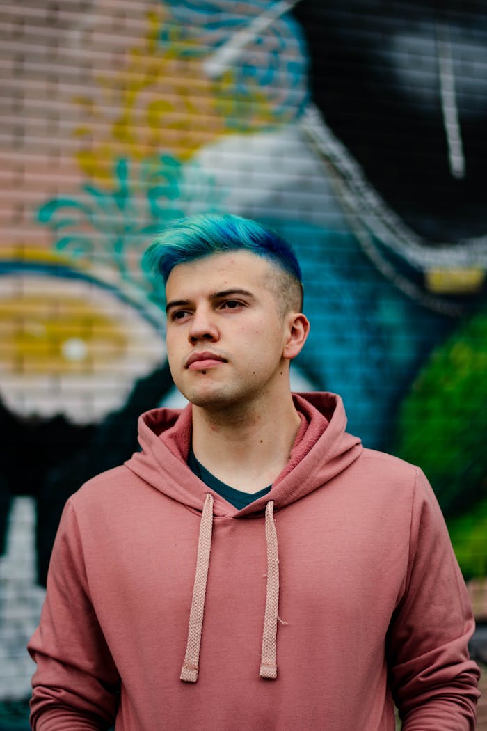 A young man in a pink hoodie with blue hair stands against a vibrant graffiti wall.