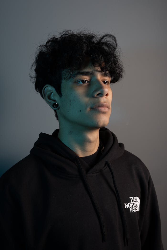Portrait of a young man wearing a black hoodie with a piercing, set against a gray studio background.